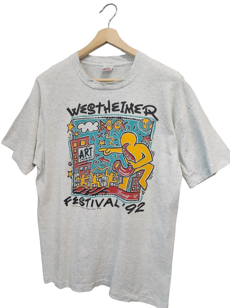 May include: A light gray t-shirt featuring a colorful graphic for the Westheimer Art Festival '92. The design includes a yellow figure playing a saxophone, buildings, and the word "ART". The shirt is hanging on a wooden hanger.