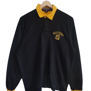 Könnte beinhalten: Schwarzes Rugby-Langarmshirt mit gelbem Kragen und Bündchen. Das Shirt hat den Text "POLO SPORT" und ein Logo auf der Brust. Das Shirt hängt an einem Holzkleiderbügel.