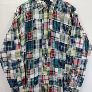 May include: A long-sleeved patchwork shirt in a variety of plaid patterns, including red, green, blue, and white. The shirt has a button-down collar and a small embroidered logo on the chest. The shirt is hanging on a wooden hanger.
