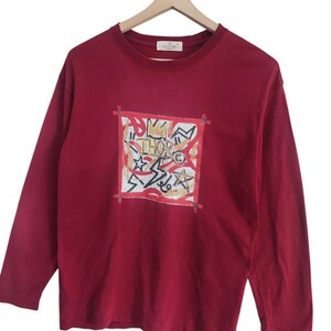 Puede incluir: Una camiseta de manga larga de color burdeos con un dise&ntilde;o gr&aacute;fico en la parte delantera. El dise&ntilde;o presenta un marco cuadrado con formas abstractas, estrellas y la palabra "THOR" en el centro. La camiseta est&aacute; colgada en una percha de madera.