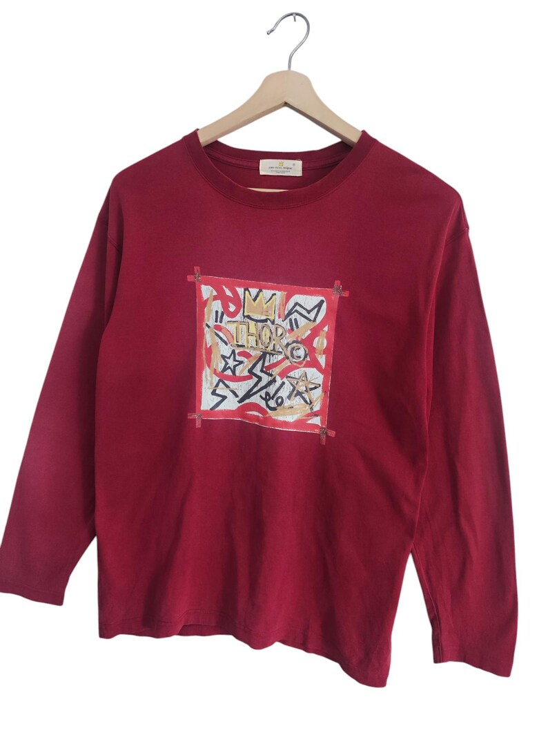 Puede incluir: Una camiseta de manga larga de color burdeos con un dise&ntilde;o gr&aacute;fico en la parte delantera. El dise&ntilde;o presenta un marco cuadrado con formas abstractas, estrellas y la palabra "THOR" en el centro. La camiseta est&aacute; colgada en una percha de madera.