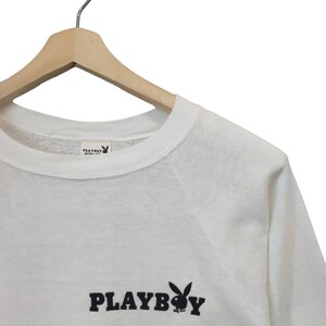 May include: White sweatshirt with the word "PLAYBOY" in black block letters and a black bunny logo. The sweatshirt is hanging on a wooden hanger.