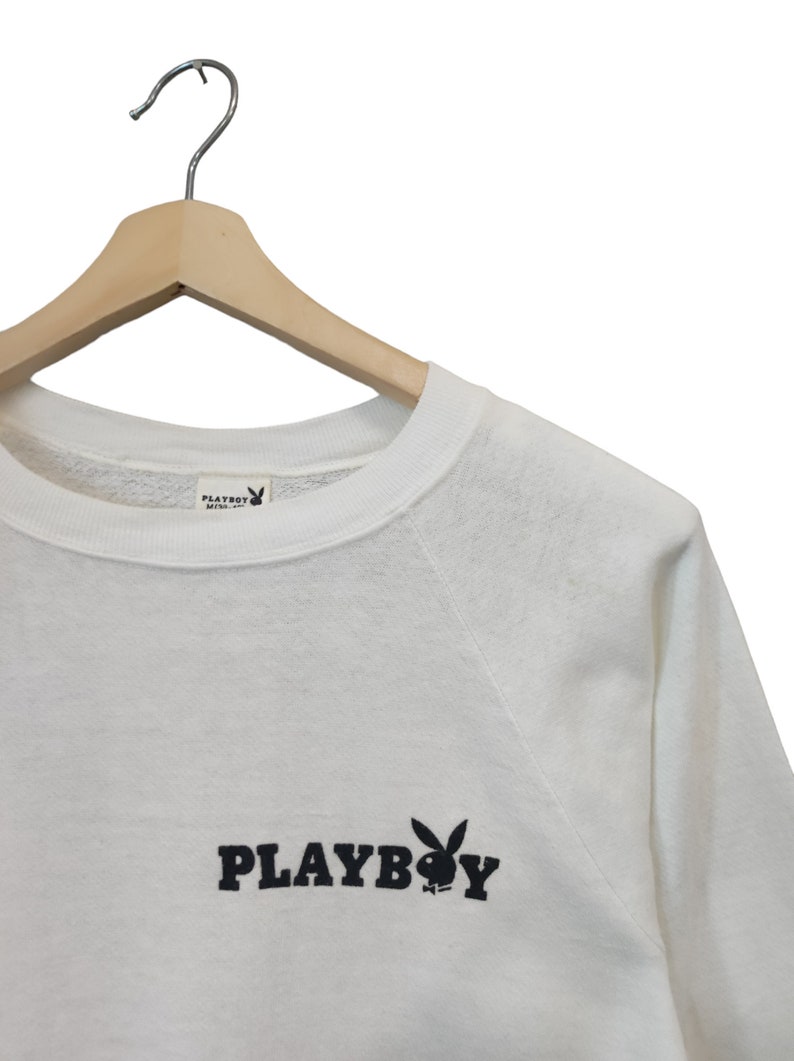 May include: White sweatshirt with the word "PLAYBOY" in black block letters and a black bunny logo. The sweatshirt is hanging on a wooden hanger.