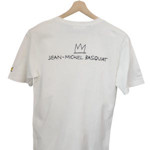 Puede incluir: Camiseta blanca con la inscripci&oacute;n "JEAN-MICHEL BASQUIAT" y un dise&ntilde;o de corona negra. La camiseta est&aacute; colgada en una percha de madera sobre un fondo blanco. La camiseta tiene mangas cortas.
