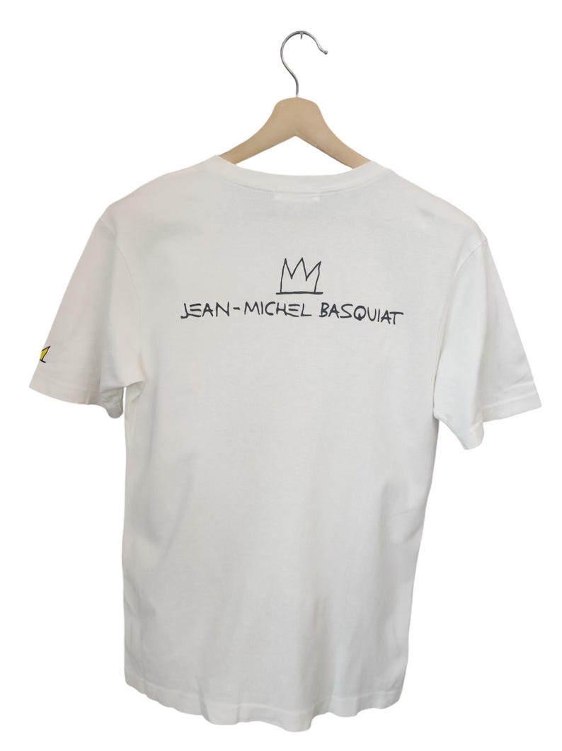 Puede incluir: Camiseta blanca con la inscripci&oacute;n "JEAN-MICHEL BASQUIAT" y un dise&ntilde;o de corona negra. La camiseta est&aacute; colgada en una percha de madera sobre un fondo blanco. La camiseta tiene mangas cortas.