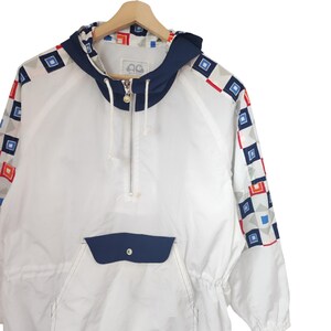 May include: White anorak jacket with a navy blue hood, pocket, and trim. The sleeves feature a geometric pattern of red, blue, and gray squares. The jacket has a half-zip closure and a drawstring at the waist.
