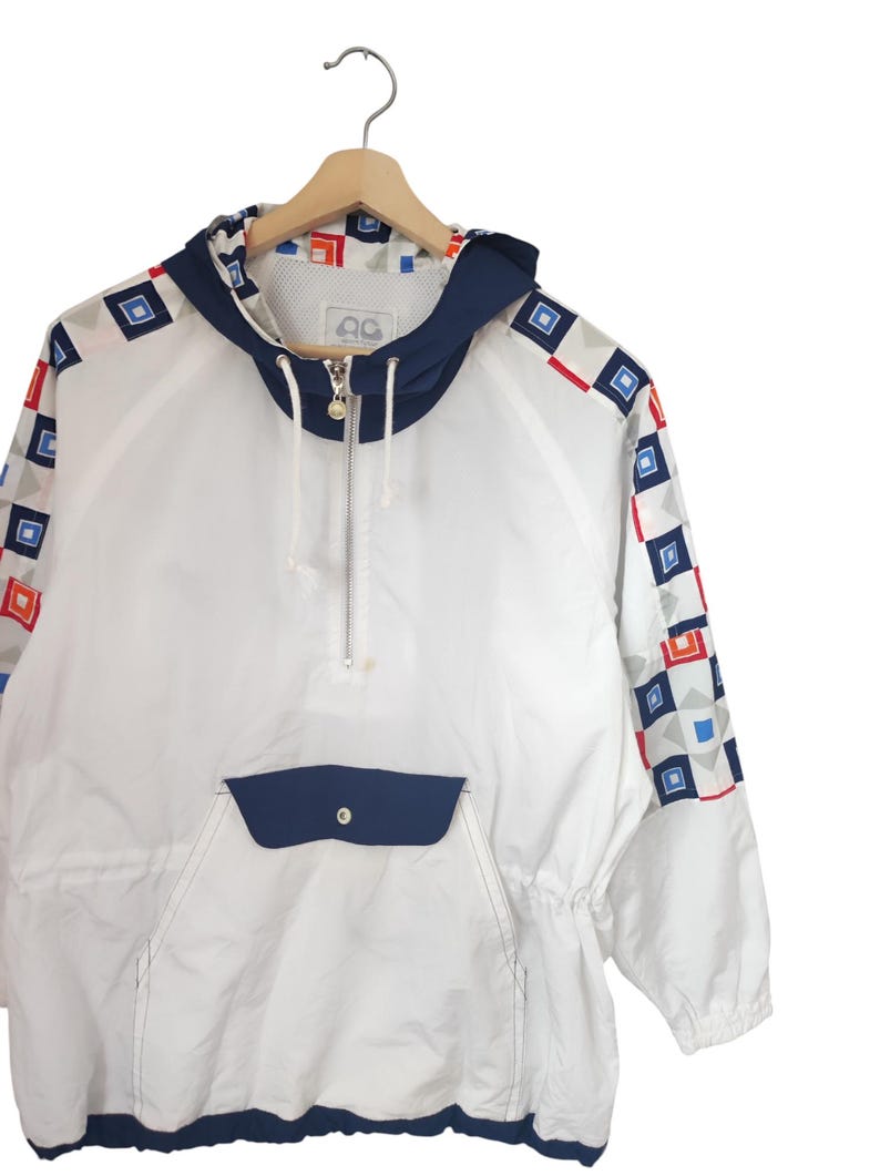 May include: White anorak jacket with a navy blue hood, pocket, and trim. The sleeves feature a geometric pattern of red, blue, and gray squares. The jacket has a half-zip closure and a drawstring at the waist.