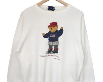 May include: Off-white sweatshirt featuring a Polo Bear graphic. The bear wears a red cap, a navy blue sweater with an American flag, and a gray skirt. The text "POLO BEAR by RALPH LAUREN" is printed below the bear.