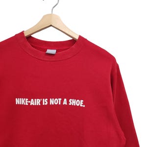 May include: A red crew neck sweatshirt with the text "NIKE-AIR IS NOT A SHOE" in white. The sweatshirt is hanging on a wooden hanger with a metal hook.