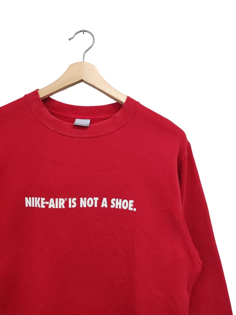 May include: A red crew neck sweatshirt with the text "NIKE-AIR IS NOT A SHOE" in white. The sweatshirt is hanging on a wooden hanger with a metal hook.