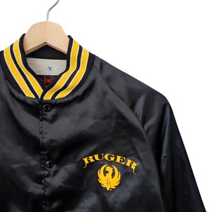 May include: Black satin jacket with a yellow and black striped collar. The jacket features the word "RUGER" in gold lettering with a gold emblem. The jacket is hanging on a wooden hanger.