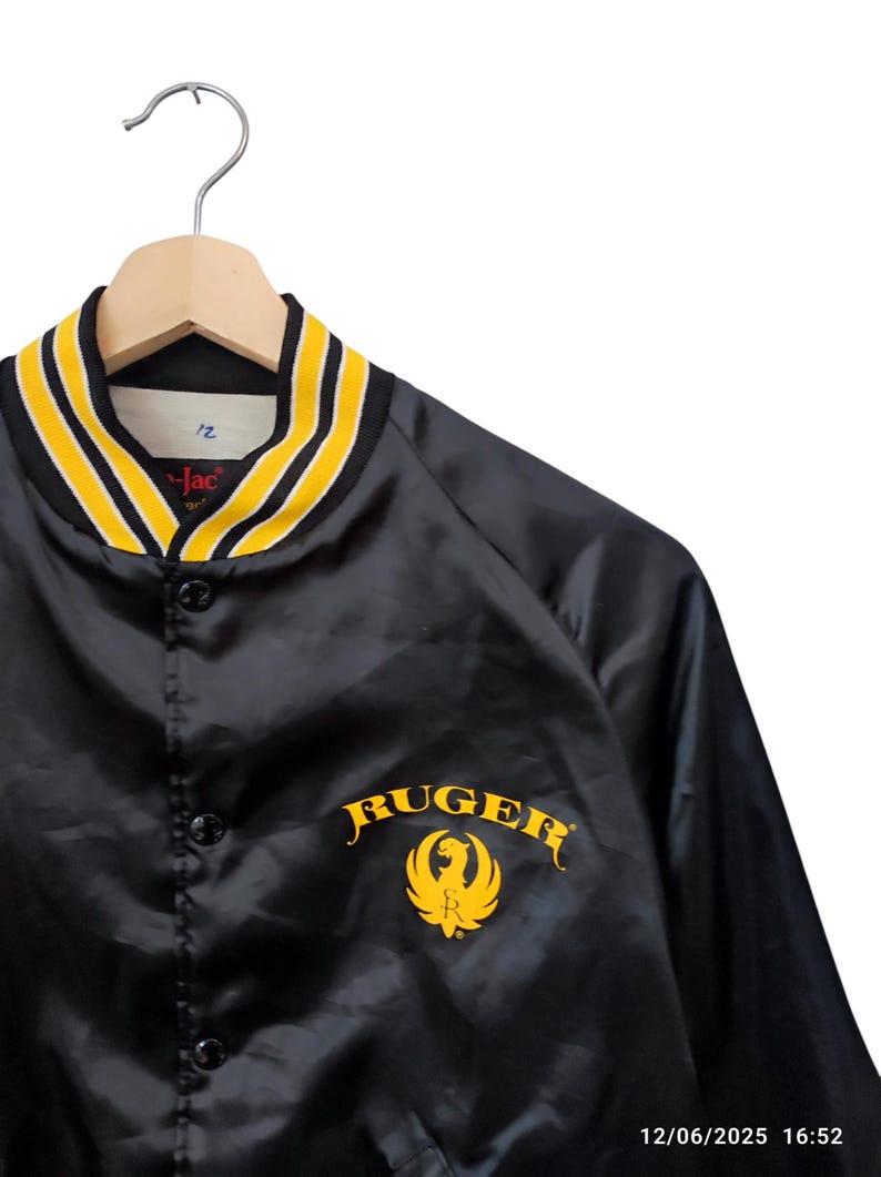 May include: Black satin jacket with a yellow and black striped collar. The jacket features the word "RUGER" in gold lettering with a gold emblem. The jacket is hanging on a wooden hanger.