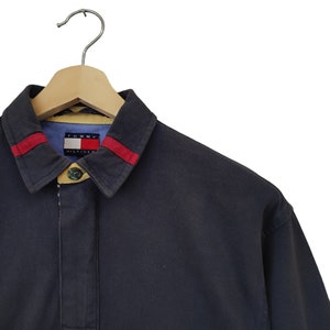 May include: Navy blue long-sleeve shirt with a collar and red stripes. The shirt has a buttoned placket and a Tommy Hilfiger logo on the inside collar. The shirt is hanging on a wooden hanger.