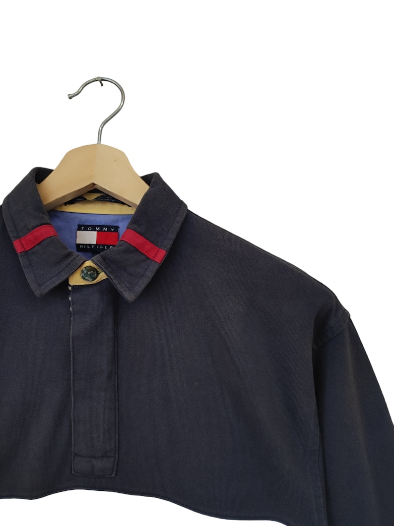 May include: Navy blue long-sleeve shirt with a collar and red stripes. The shirt has a buttoned placket and a Tommy Hilfiger logo on the inside collar. The shirt is hanging on a wooden hanger.