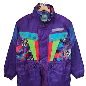 May include: A vintage purple ski jacket with blue and teal accents. The jacket features a high collar, multiple pockets, and a colorful geometric pattern on the front. The text "CCLTEAM" is visible on the jacket.