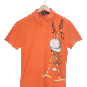 Puede incluir: Polo naranja con un diseño de polo. El diseño incluye palos de polo, un casco y una pelota. La camisa tiene un cuello clásico y una tapeta de botones. Cuelga de una percha de madera.