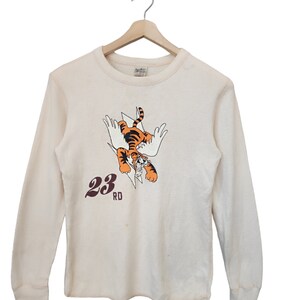 Puede incluir: Camiseta de manga larga color crema con un gráfico de un tigre alado y el texto "23 RD". El tigre es naranja y negro, sobre un fondo blanco. La camiseta tiene una textura de gofre.
