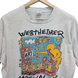May include: Heather gray t-shirt with a colorful graphic from the Westheimer Festival '92. The design includes a figure playing a saxophone, buildings, stars, and the word "ART". The shirt is hanging on a wooden hanger.