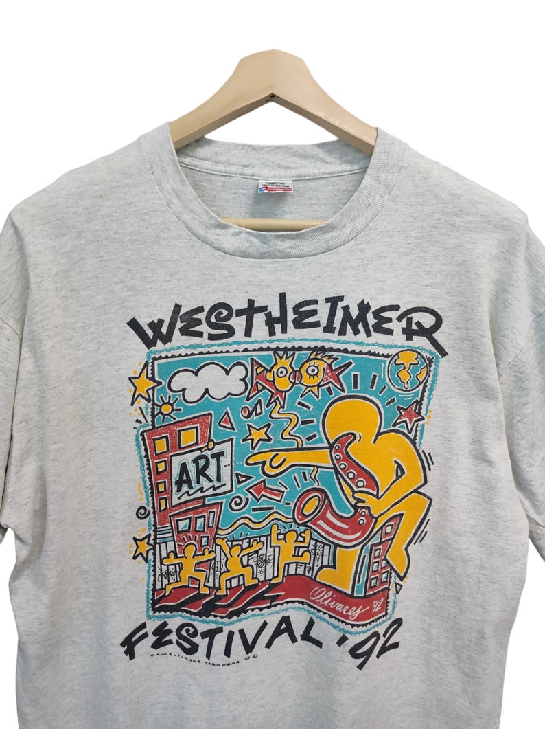May include: Heather gray t-shirt with a colorful graphic from the Westheimer Festival '92. The design includes a figure playing a saxophone, buildings, stars, and the word "ART". The shirt is hanging on a wooden hanger.