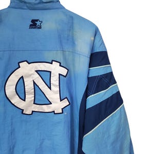 May include: A light blue and navy blue Starter windbreaker jacket featuring a large white and navy blue embroidered NC logo on the back. The jacket has a high collar, long sleeves with diagonal colour-blocked panels, and a Starter brand logo.