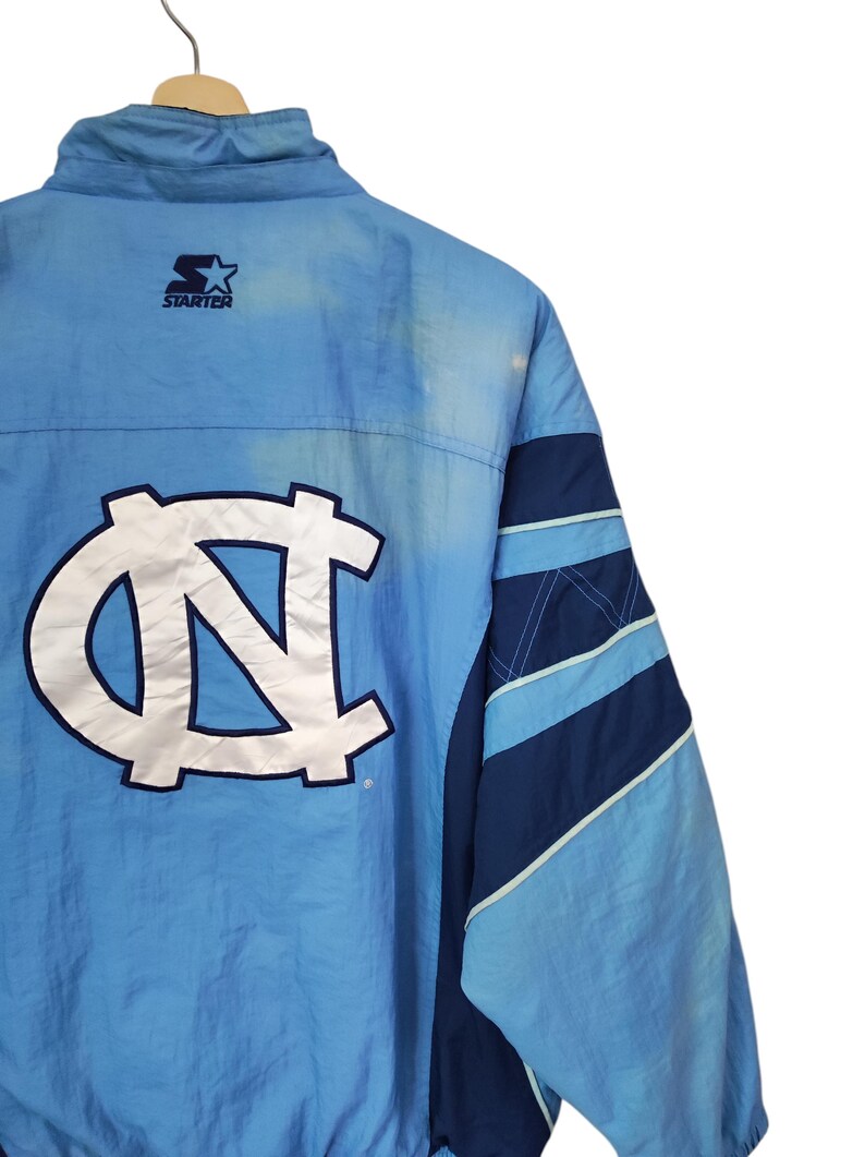 May include: A light blue and navy blue Starter windbreaker jacket featuring a large white and navy blue embroidered NC logo on the back. The jacket has a high collar, long sleeves with diagonal colour-blocked panels, and a Starter brand logo.