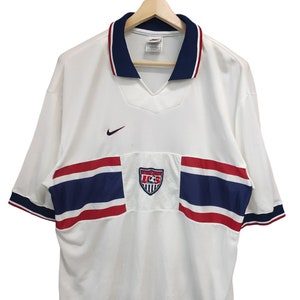 May include: White football shirt with navy and red detailing. Includes a v-neck collar, Nike logo, and a US Soccer emblem. Short-sleeved with navy and red trim. The shirt is displayed on a wooden hanger.
