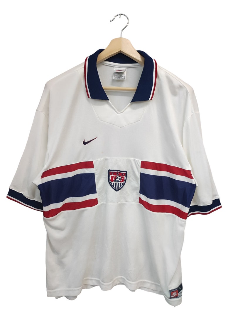 May include: White football shirt with navy and red detailing. Includes a v-neck collar, Nike logo, and a US Soccer emblem. Short-sleeved with navy and red trim. The shirt is displayed on a wooden hanger.