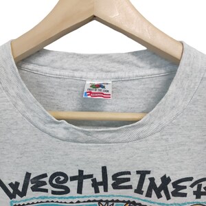May include: Light gray t-shirt with a colorful graphic design and the word "WESTHEIMER" in black. The graphic includes cartoon-like figures, stars, and a globe. The shirt is hanging on a wooden hanger.