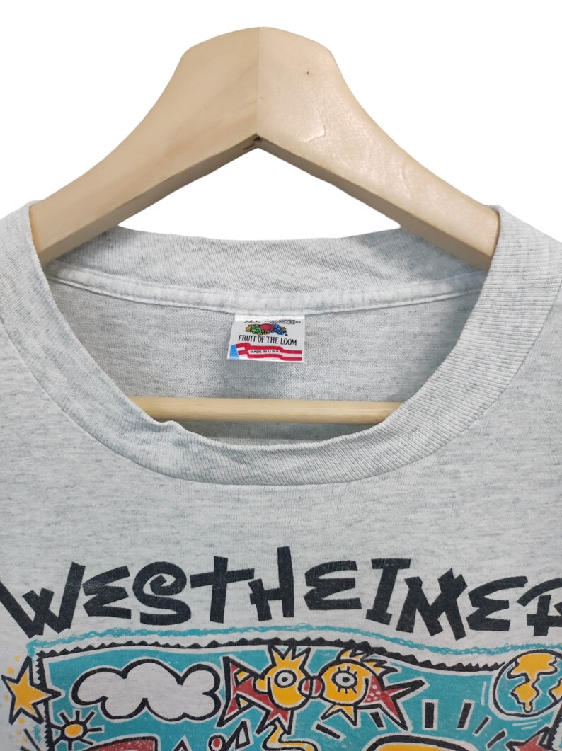 May include: Light gray t-shirt with a colorful graphic design and the word "WESTHEIMER" in black. The graphic includes cartoon-like figures, stars, and a globe. The shirt is hanging on a wooden hanger.