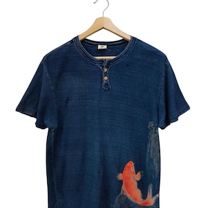 May include: Blue short-sleeved Henley shirt with a round neckline and three wooden buttons. The shirt features a vibrant orange koi fish design on the front. The shirt is hanging on a wooden hanger.