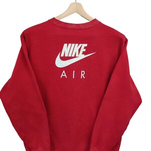 May include: Red Nike Air sweatshirt with white logo and text on the back. The sweatshirt is hanging on a wooden hanger. The Nike logo is a white swoosh above the word "NIKE" and the word "AIR".