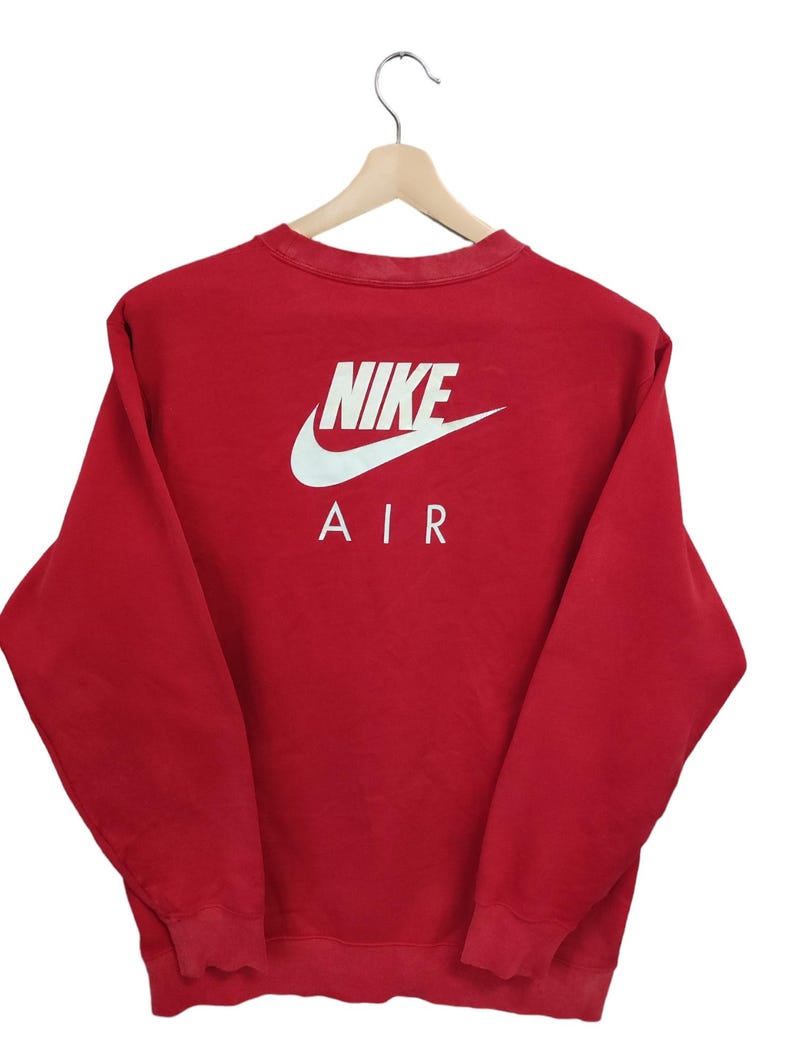 May include: Red Nike Air sweatshirt with white logo and text on the back. The sweatshirt is hanging on a wooden hanger. The Nike logo is a white swoosh above the word "NIKE" and the word "AIR".