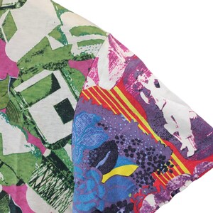 Może przedstawiać: Fragment koszulki z kr&oacute;tkim rękawem o żywym, abstrakcyjnym wzorze. Koszulka ma mieszankę kolor&oacute;w, w tym zielony, r&oacute;żowy, fioletowy, niebieski i ż&oacute;łty. Wz&oacute;r zawiera kształty kwiatowe i geometryczne.