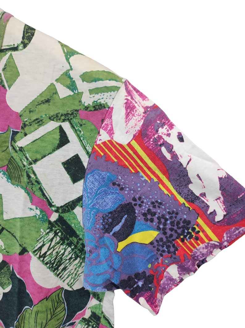 Może przedstawiać: Fragment koszulki z kr&oacute;tkim rękawem o żywym, abstrakcyjnym wzorze. Koszulka ma mieszankę kolor&oacute;w, w tym zielony, r&oacute;żowy, fioletowy, niebieski i ż&oacute;łty. Wz&oacute;r zawiera kształty kwiatowe i geometryczne.