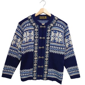 Könnte beinhalten: Marineblauer Cardigan mit traditionellem nordischem Muster in Weiß und Hellblau. Der Pullover hat einen runden Ausschnitt, lange Ärmel und Metallverschlüsse. Er wird auf einem Holzbügel präsentiert. Das Muster enthält Schneeflockenmotive.