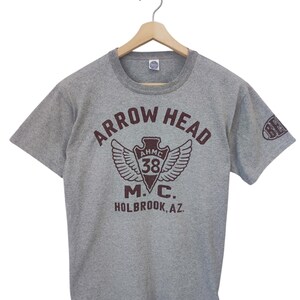 May include: A gray t-shirt with a maroon graphic that reads "ARROW HEAD M.C. HOLBROOK, AZ." The graphic includes wings, the number 38, and the letters AHMC. The shirt is hanging on a wooden hanger.