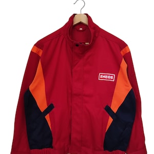 May include: A red jacket featuring a high collar, a front zipper closure, and an elasticized hem. The design includes orange diagonal panels on the chest and dark blue side panels. The word ENEOS is printed in white on a rectangular patch.