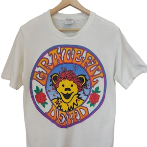 May include: White t-shirt featuring a circular graphic with the words "Grateful Dead" in orange and purple. The graphic includes a yellow bear wearing a floral crown and surrounded by roses. The shirt is hanging on a wooden hanger.
