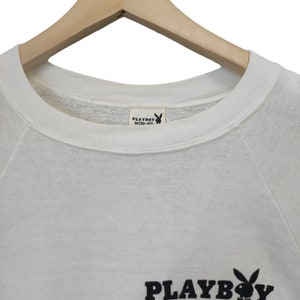 May include: White sweatshirt with the Playboy logo in black. The logo features the word "PLAYBOY" and the iconic bunny silhouette. The sweatshirt is hanging on a wooden hanger.