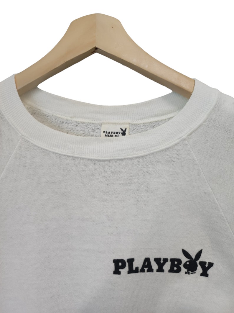 May include: White sweatshirt with the Playboy logo in black. The logo features the word "PLAYBOY" and the iconic bunny silhouette. The sweatshirt is hanging on a wooden hanger.