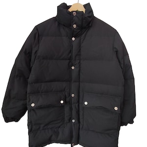 May include: A black puffer jacket with a high collar and snap closures. The jacket has two front pockets with snap closures and is hanging on a wooden hanger. The jacket is a winter outerwear item.