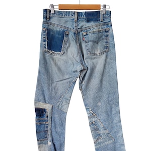 Puede incluir: Vaqueros de mezclilla azul claro con múltiples parches y detalles desgastados. Los vaqueros cuelgan de una percha de madera. Los vaqueros tienen una etiqueta roja de Levi's en el bolsillo trasero.