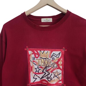 Puede incluir: Una sudadera burdeos con un dise&ntilde;o gr&aacute;fico. El dise&ntilde;o presenta la palabra "THOR" con una corona, estrellas y formas abstractas en rojo, blanco y dorado. La sudadera est&aacute; colgada en una percha de madera.