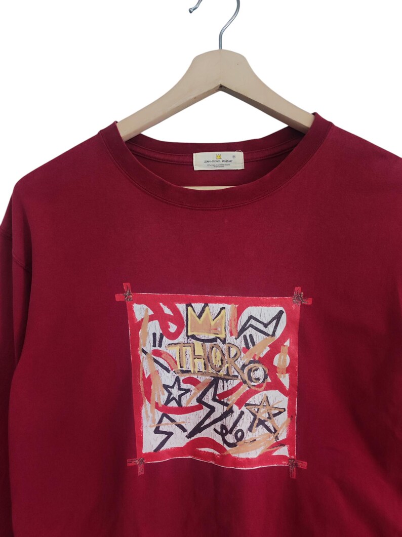 Puede incluir: Una sudadera burdeos con un dise&ntilde;o gr&aacute;fico. El dise&ntilde;o presenta la palabra "THOR" con una corona, estrellas y formas abstractas en rojo, blanco y dorado. La sudadera est&aacute; colgada en una percha de madera.