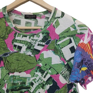 Może przedstawiać: T-shirt z kr&oacute;tkim rękawem z żywym, abstrakcyjnym wzorem kwiatowym w kolorze zielonym, białym i r&oacute;żowym. Rękaw po prawej stronie ma kolorowy, mieszany wz&oacute;r. Koszulka wisi na drewnianym wieszaku.