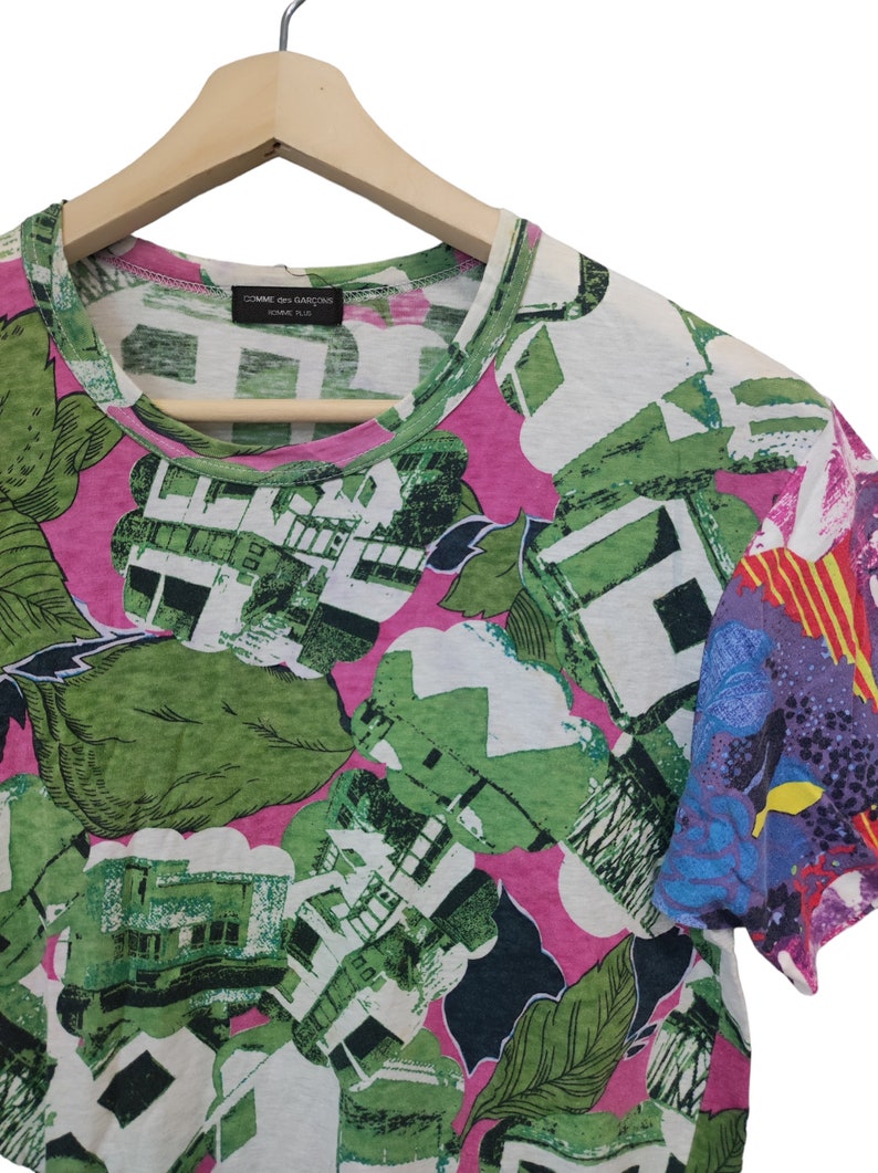 Może przedstawiać: T-shirt z kr&oacute;tkim rękawem z żywym, abstrakcyjnym wzorem kwiatowym w kolorze zielonym, białym i r&oacute;żowym. Rękaw po prawej stronie ma kolorowy, mieszany wz&oacute;r. Koszulka wisi na drewnianym wieszaku.