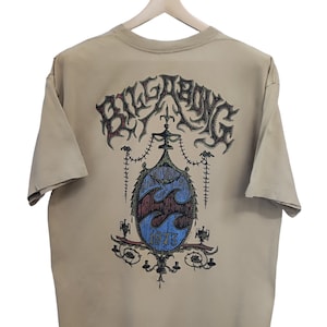 May include: A tan t-shirt with a graphic design. The design includes the word "Billabong" above a crest with waves and the year "1973". The t-shirt is hanging on a wooden hanger.