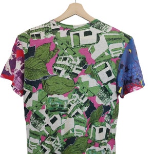 Może przedstawiać: T-shirt z kr&oacute;tkim rękawem z żywym, całopowierzchniowym nadrukiem. Wz&oacute;r przedstawia zielone liście, białe kształty architektoniczne i r&oacute;żowe akcenty. Rękawy mają kontrastujące wzory czerwonych, fioletowych i niebieskich kwiat&oacute;w. Koszulka wisi na drewnianym wieszaku.