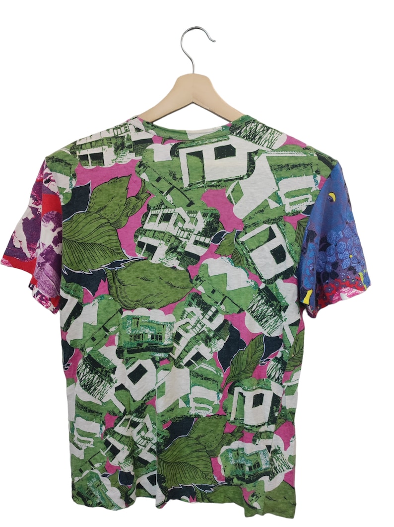 Może przedstawiać: T-shirt z kr&oacute;tkim rękawem z żywym, całopowierzchniowym nadrukiem. Wz&oacute;r przedstawia zielone liście, białe kształty architektoniczne i r&oacute;żowe akcenty. Rękawy mają kontrastujące wzory czerwonych, fioletowych i niebieskich kwiat&oacute;w. Koszulka wisi na drewnianym wieszaku.