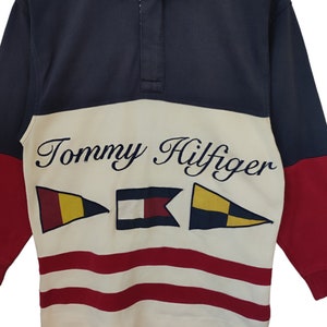 May include: A navy blue, red, and cream-colored long-sleeve shirt with the Tommy Hilfiger logo and nautical flag graphics. The shirt has a collared neckline and a buttoned placket.